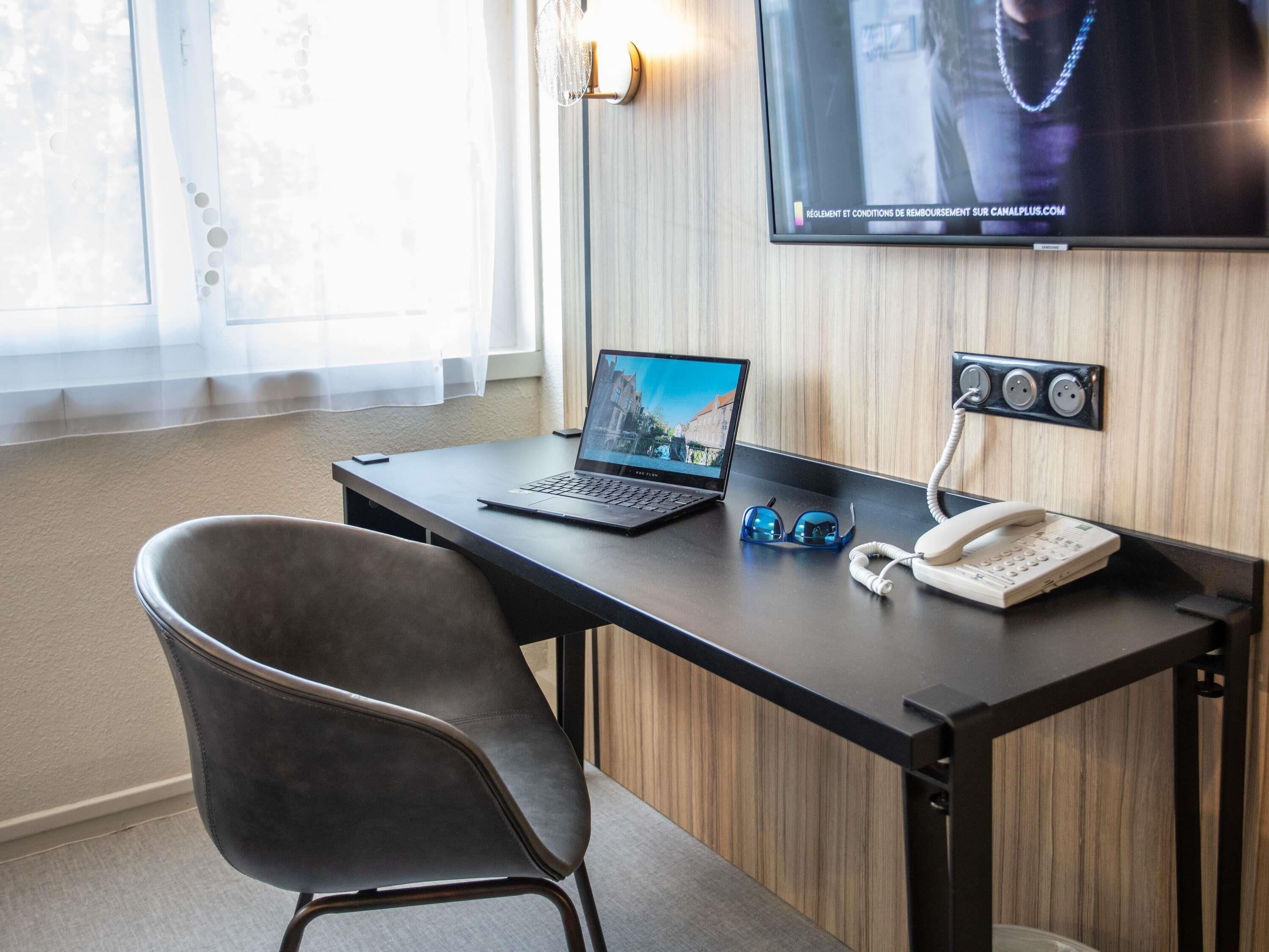 Quarto de hotel com escrivaninha, laptop, óculos, telefone e TV fixada na parede.