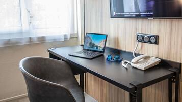 Quarto de hotel com escrivaninha, laptop, óculos, telefone e TV fixada na parede.