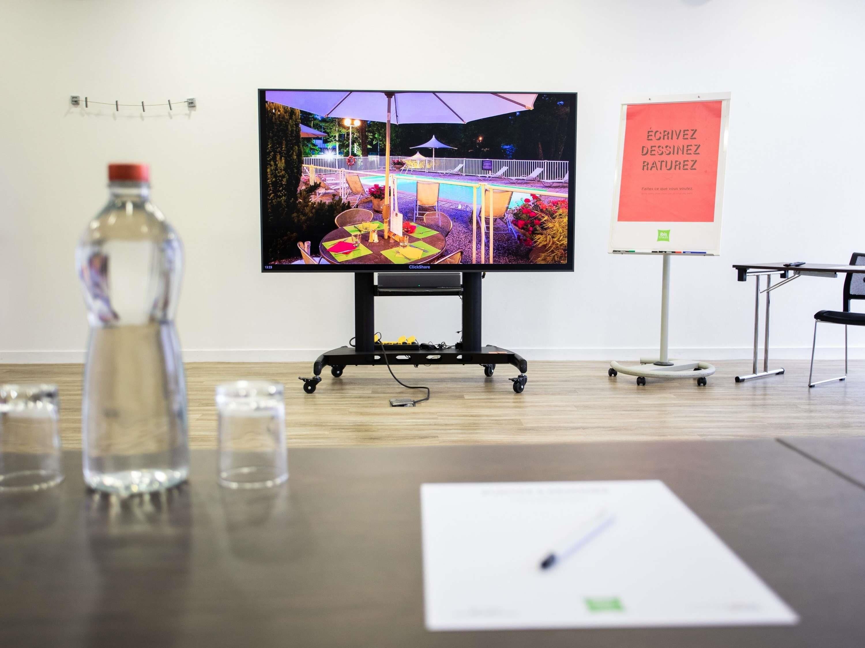 A conference room with a large screen displaying an outdoor event, a whiteboard with text, and a table with a bottle and glasses.