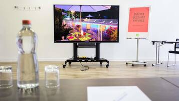 A conference room with a large screen displaying an outdoor event, a whiteboard with text, and a table with a bottle and glasses.