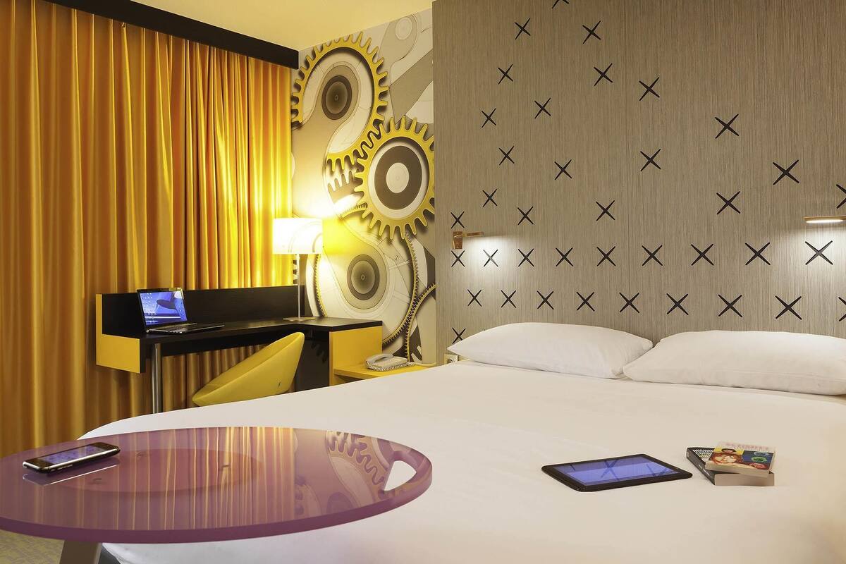 Ein Hotelzimmer mit einem Bett, einem Schreibtisch mit einem Laptop, einem runden Tisch mit einem Smartphone und einer Wand mit geometrischem Muster.
