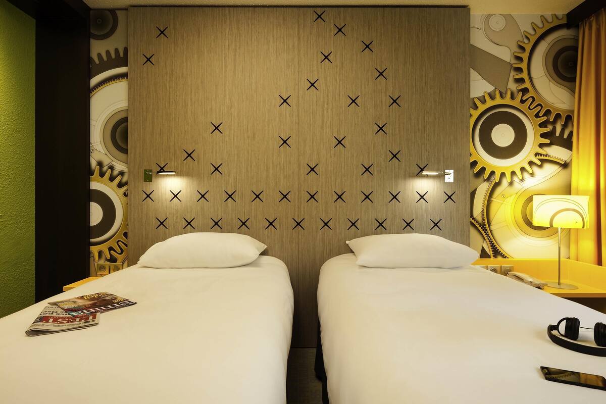 Ein Hotelzimmer mit zwei Betten, einem Holzkopfstück mit geometrischem Muster, einer Wand mit großen Zahnrädern und einer Lampe auf dem Nachttisch.
