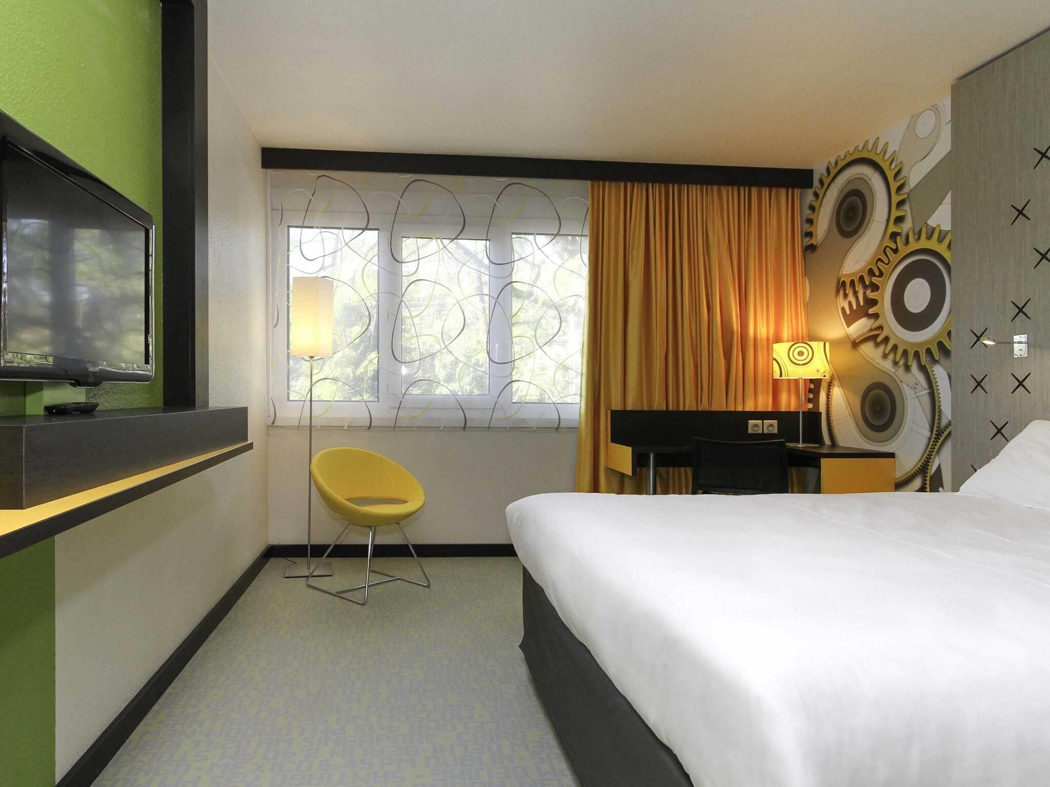 A hotel room with a bed, a TV mounted on the wall, a desk with a lamp, a chair, and a window with curtains.