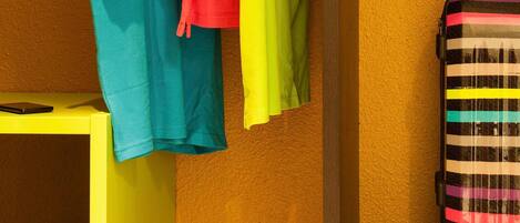A room with a yellow table, a striped suitcase, and colorful hanging clothes.