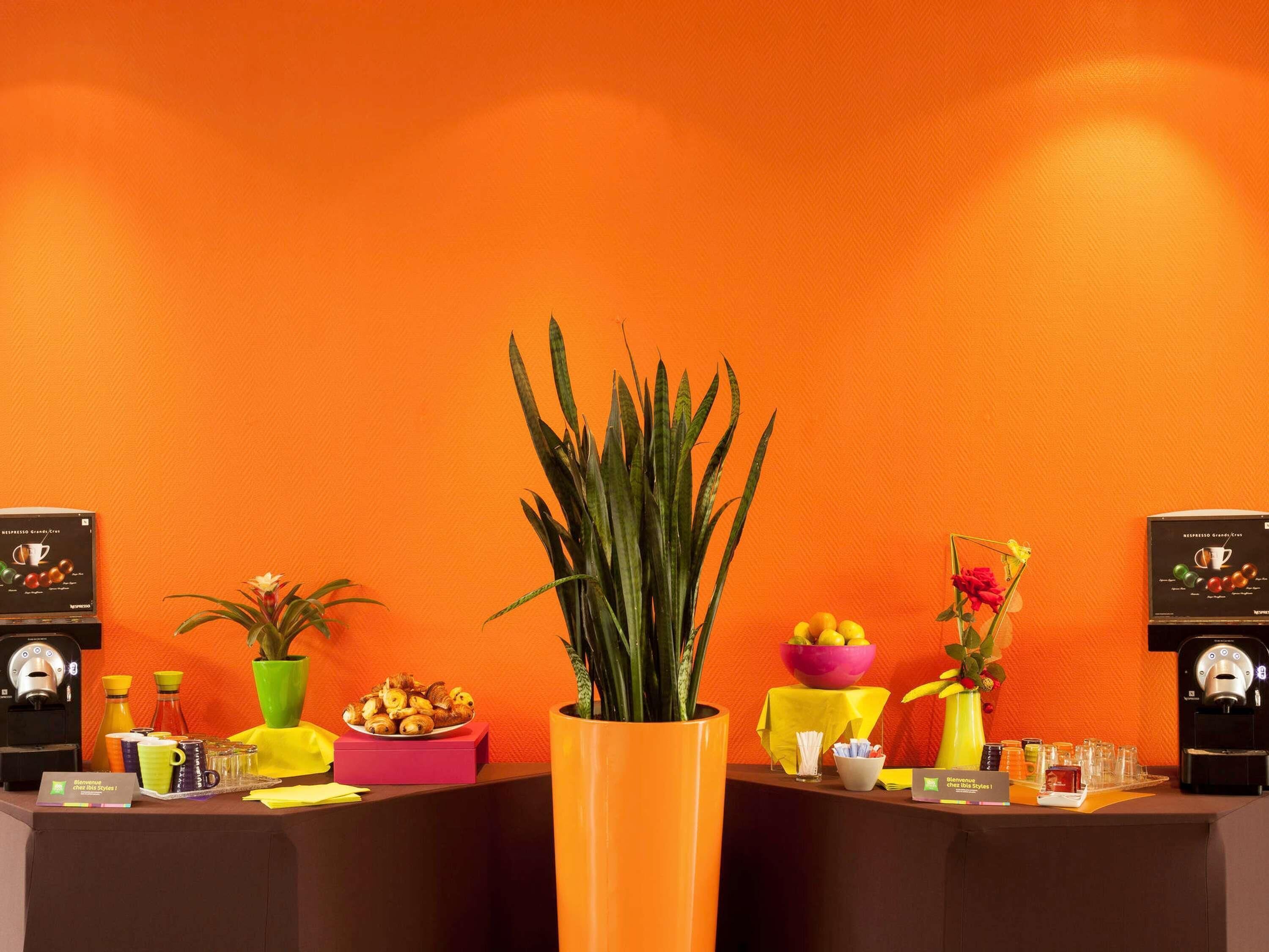 Un comptoir d’accueil avec une machine à café, un saladier de fruits, une plante en pot et un vase rempli de fleurs.