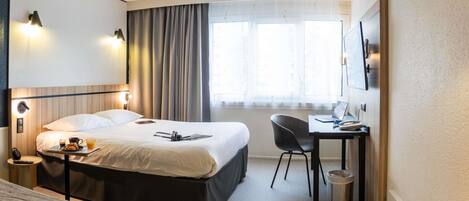 A hotel room with a bed, a desk, a chair, a bedside table with a lamp, a window with curtains, and a trash bin.