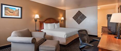A hotel room with a bed, two chairs, a desk, a TV, and a framed picture on the wall.