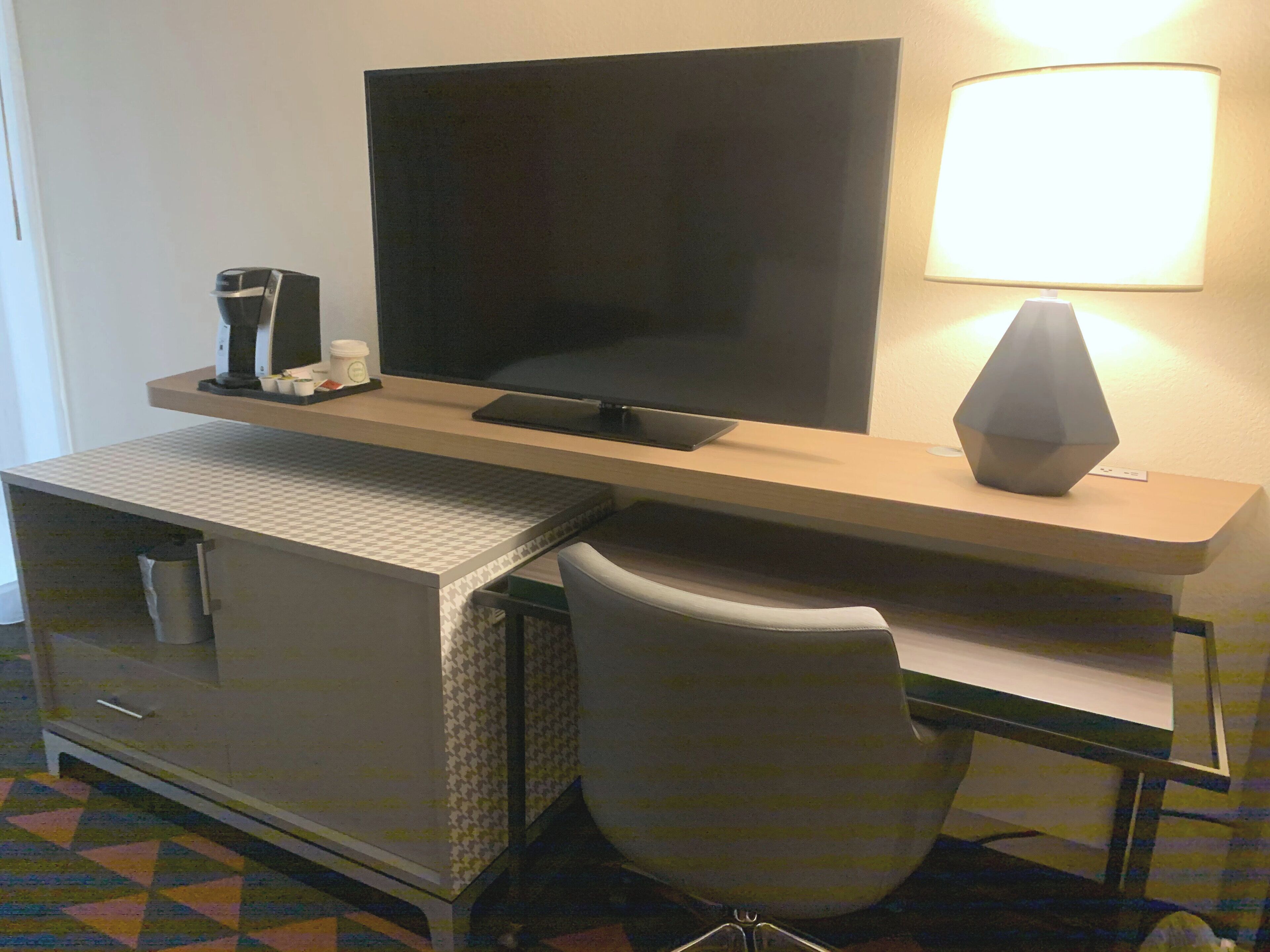 A hotel room with a flat-screen TV, a desk with a lamp, a chair, and a minibar.