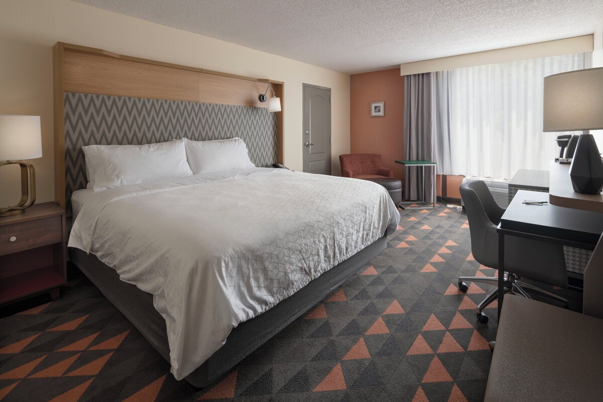 Une chambre d’hôtel avec un grand lit, un bureau, une chaise, une lampe et une fenêtre avec des rideaux.