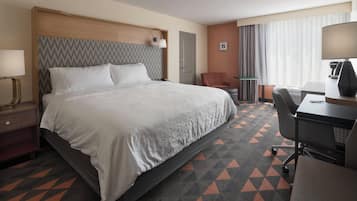 Quarto de hotel com uma cama grande, escrivaninha, cadeira, luminária e janela com cortinas.