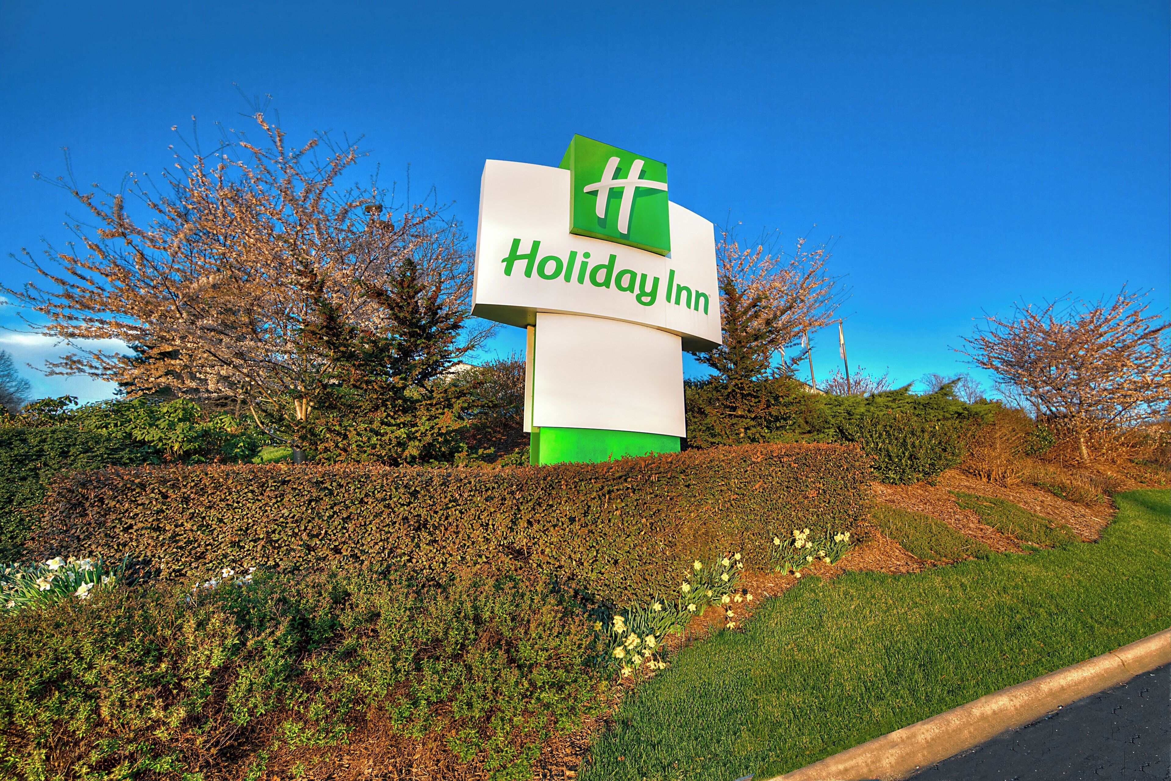 A Holiday Inn sign with a green and white logo, surrounded by well-maintained hedges and trees.