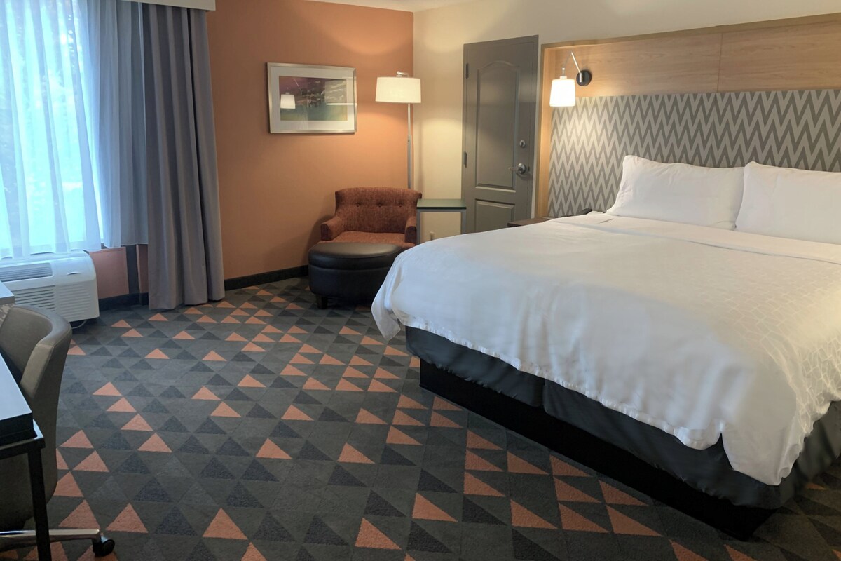 Une chambre d’hôtel avec un lit, une chaise, un bureau et une fenêtre avec des rideaux.