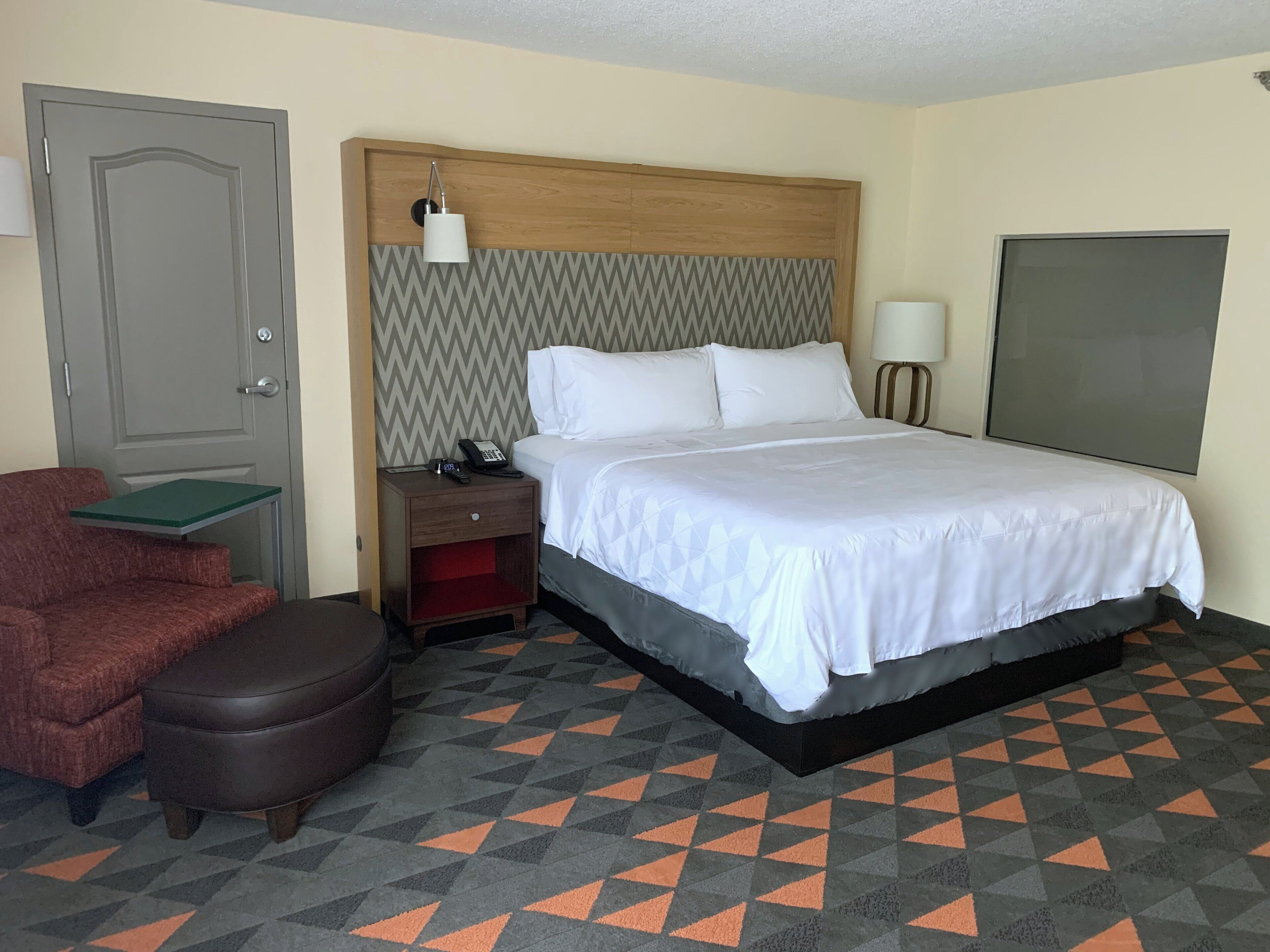Una habitación de hotel con cama, mesita de noche, teléfono, silla y puerta.
