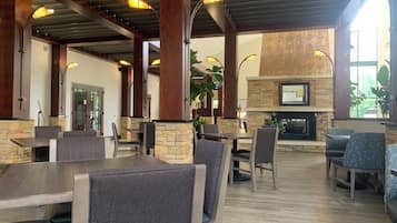 A spacious indoor area with multiple tables and chairs, a stone fireplace, and large windows allowing natural light.