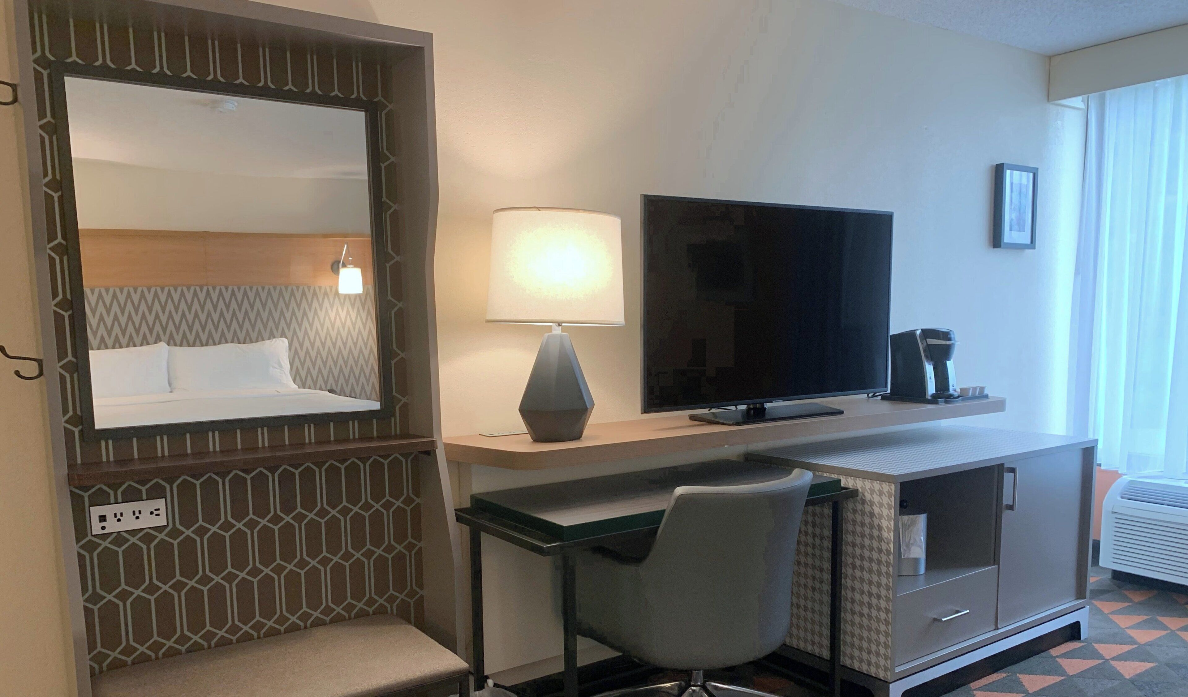 Ein Hotelzimmer mit einem Bett, einem Schreibtisch mit Stuhl, einem Fernseher und einem Fenster mit Vorhängen.