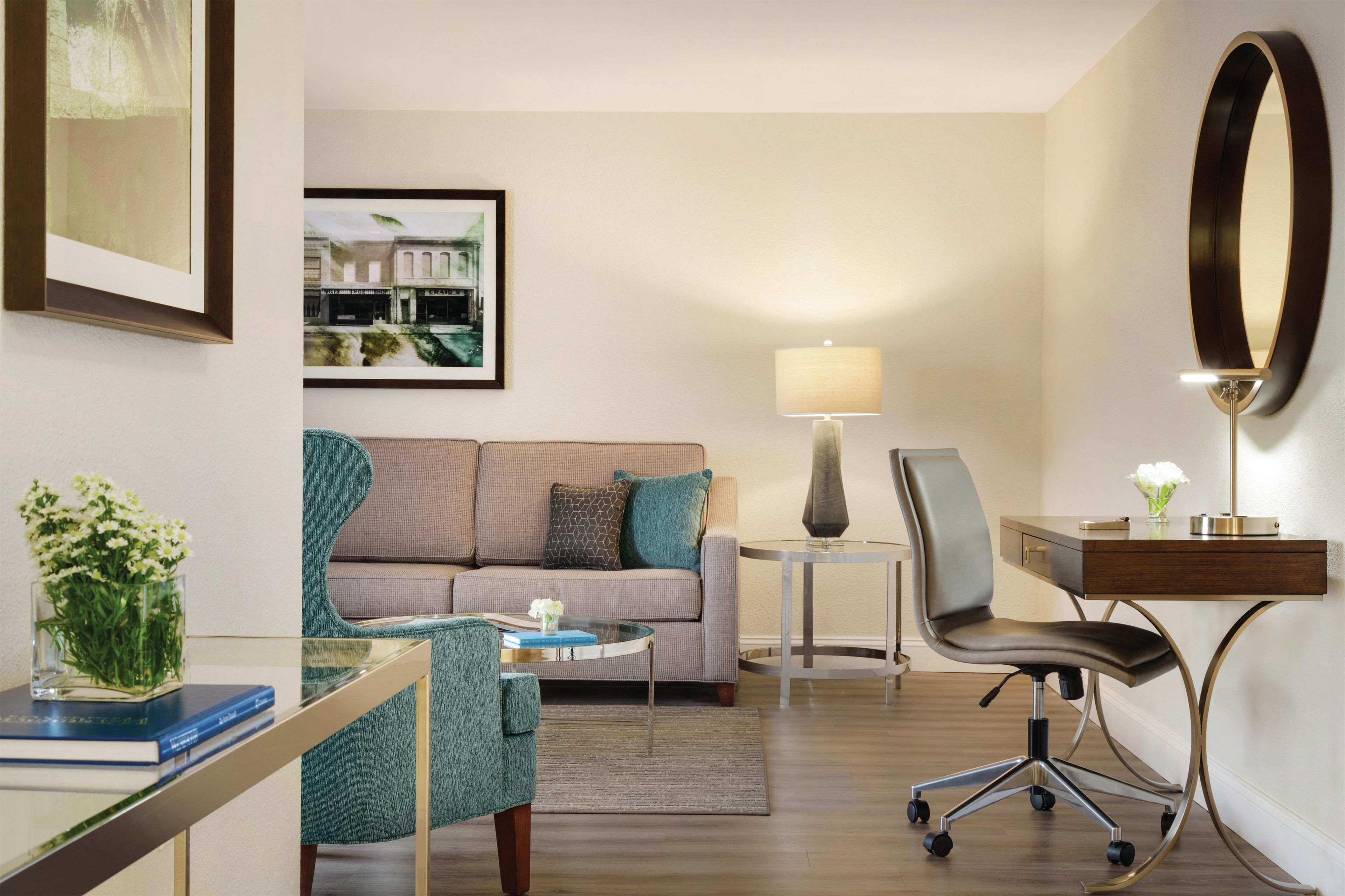 Una habitación de hotel moderna con sofá, escritorio con silla, lámpara, espejo y un cuadro en la pared.