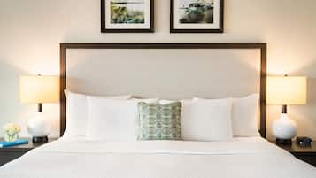 Una cama bien hecha con sábanas blancas y un cojín decorativo, flanqueada por dos lámparas de noche y una mesita con un reloj y un pequeño jarrón de flores.