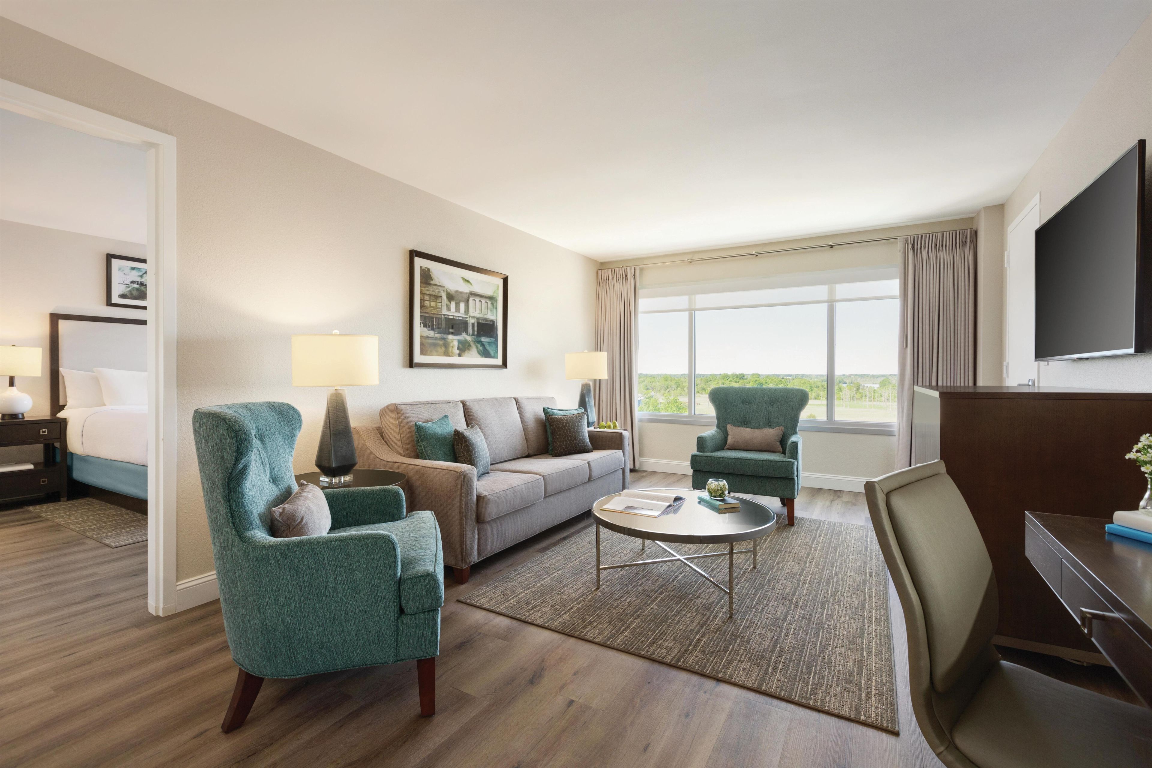 Une chambre d’hôtel comprenant un lit, un canapé, deux fauteuils, une table basse, une télévision et une grande fenêtre donnant sur un paysage agréable.