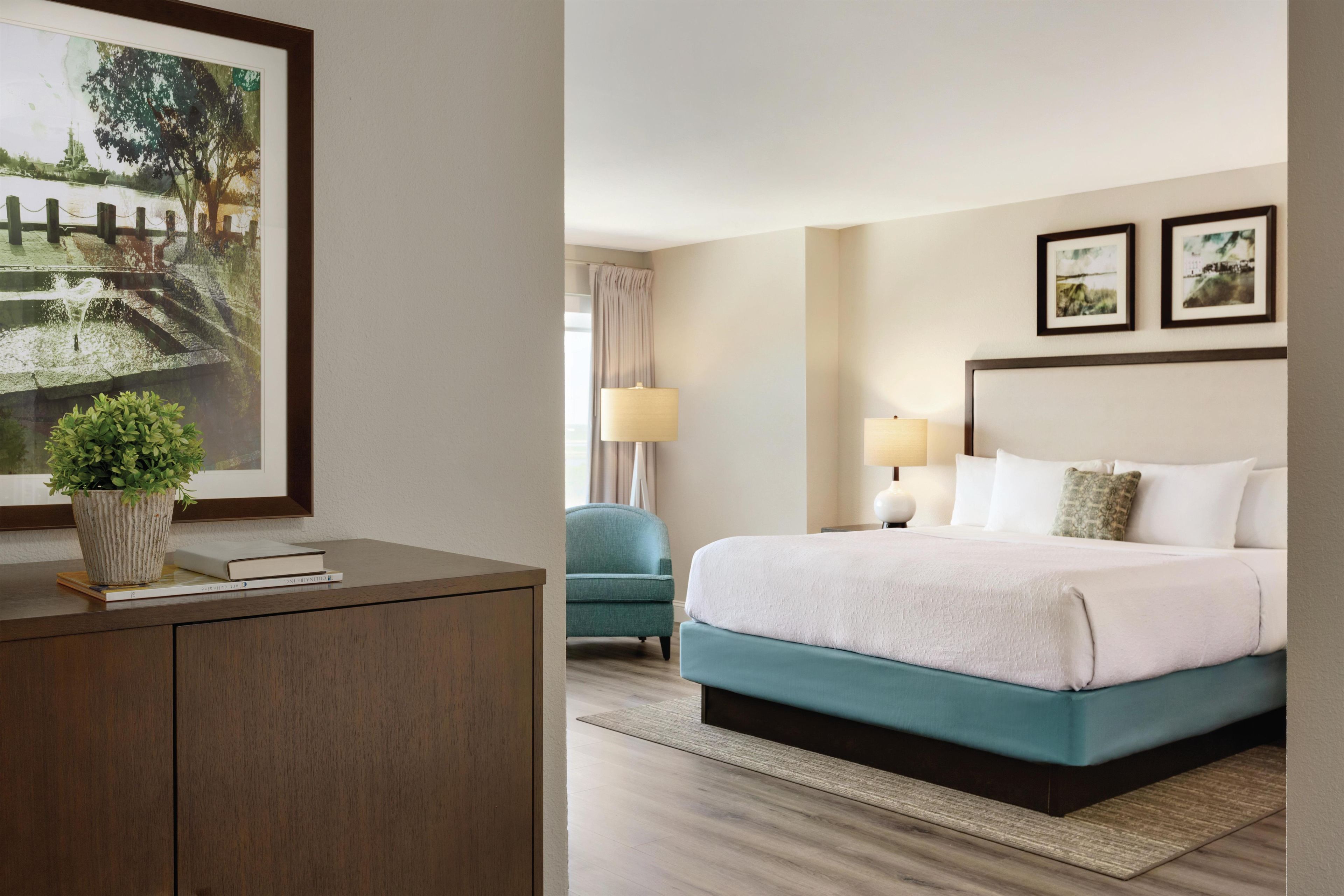 Habitación de hotel con cama, lámparas de noche, escritorio con planta y un cuadro en la pared.