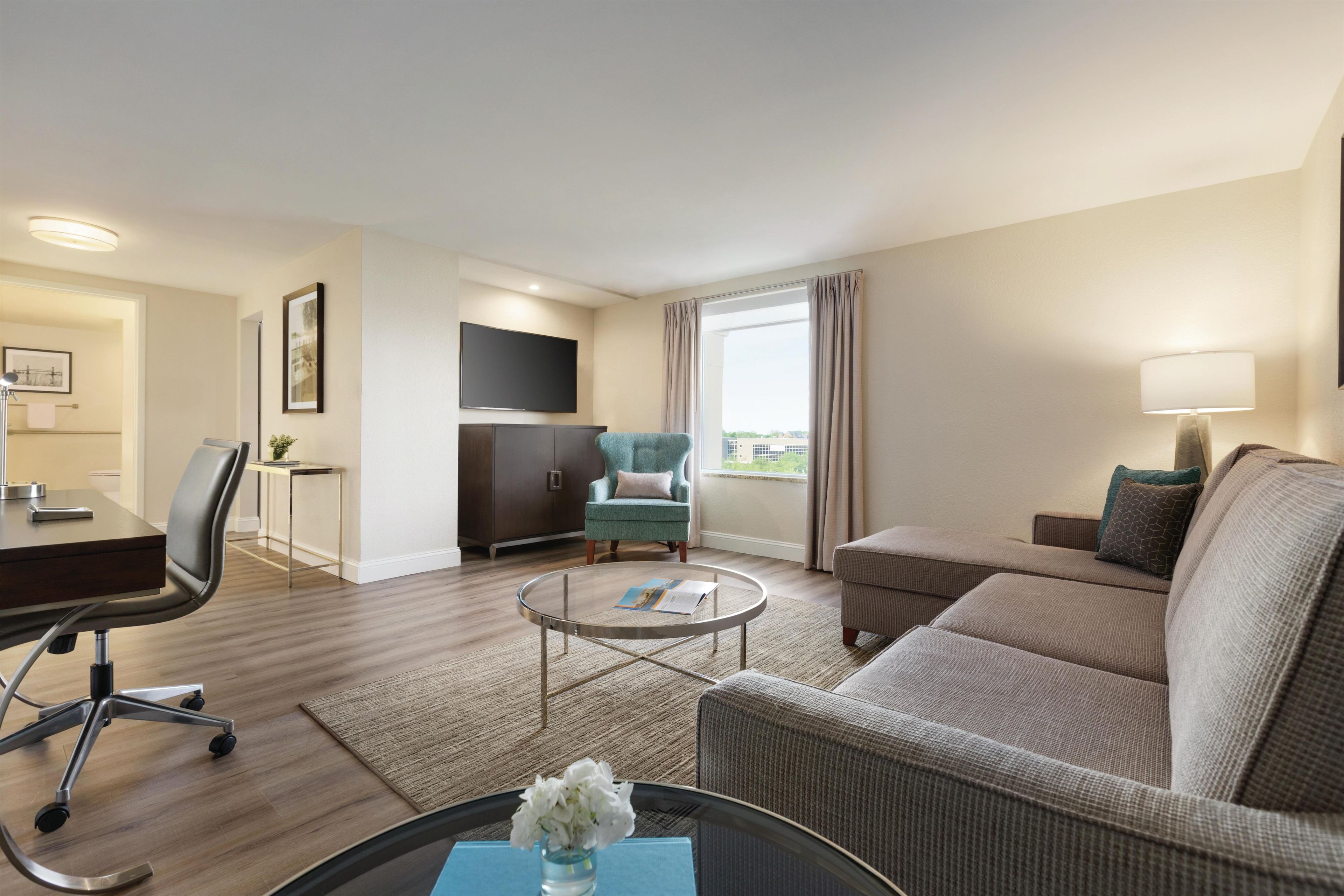 A hotel room with a sofa, armchair, TV, desk, and a view of the city.