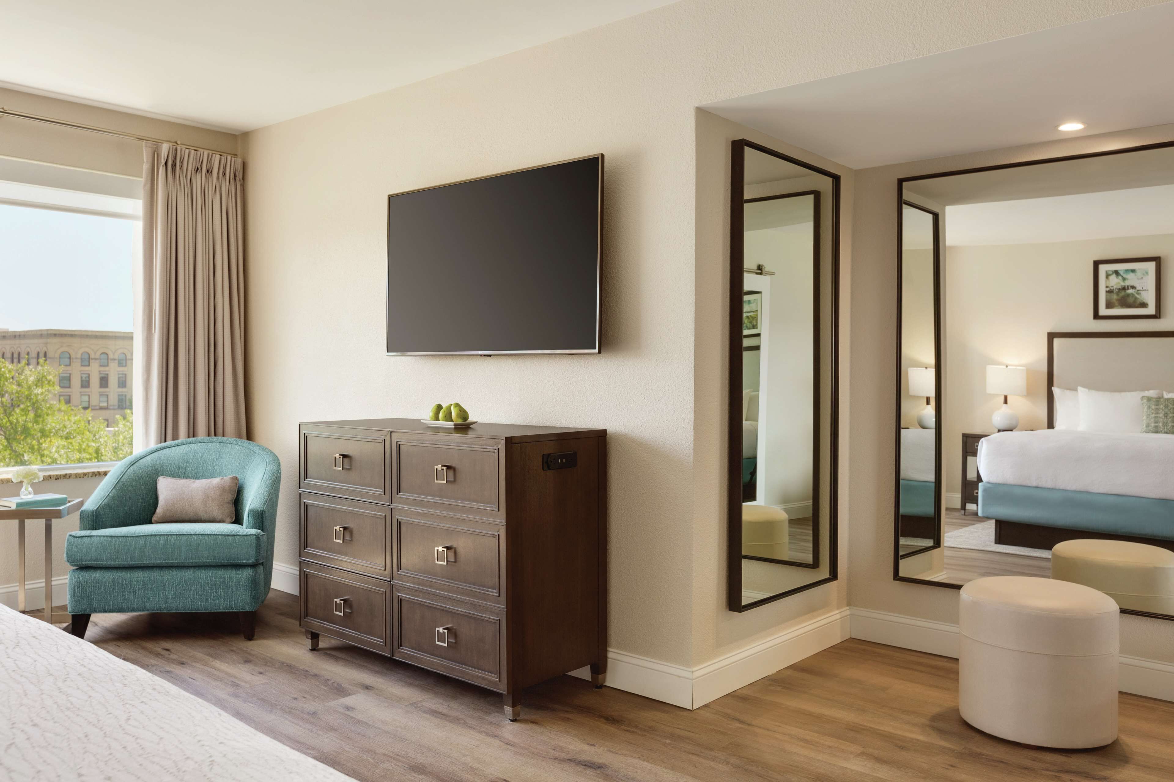 A hotel room with a large bed, a dresser, a chair, a flat-screen TV, a large mirror, and a window with curtains.