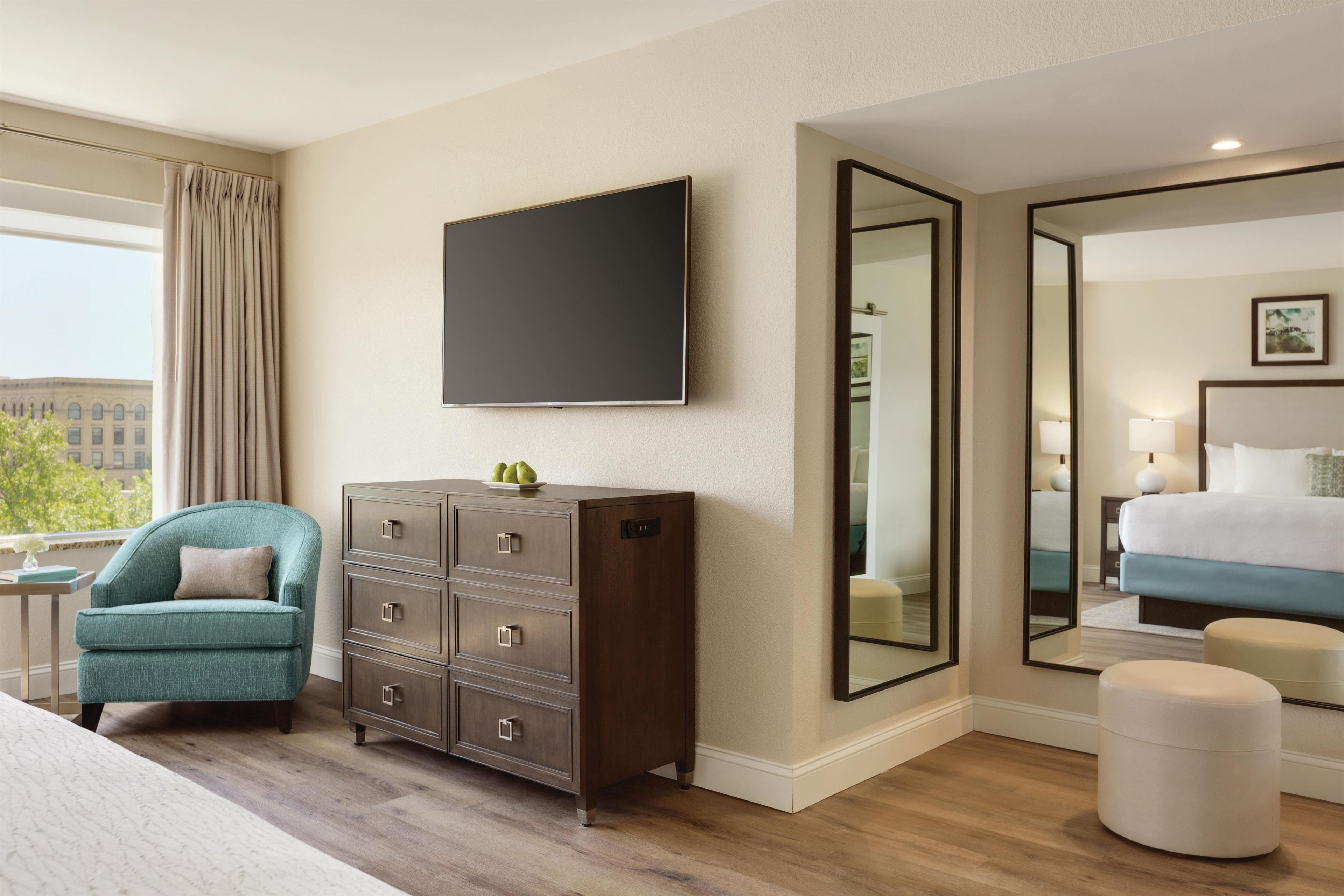 A hotel room with a bed, a dresser, a television, a large mirror, and a view of the city.