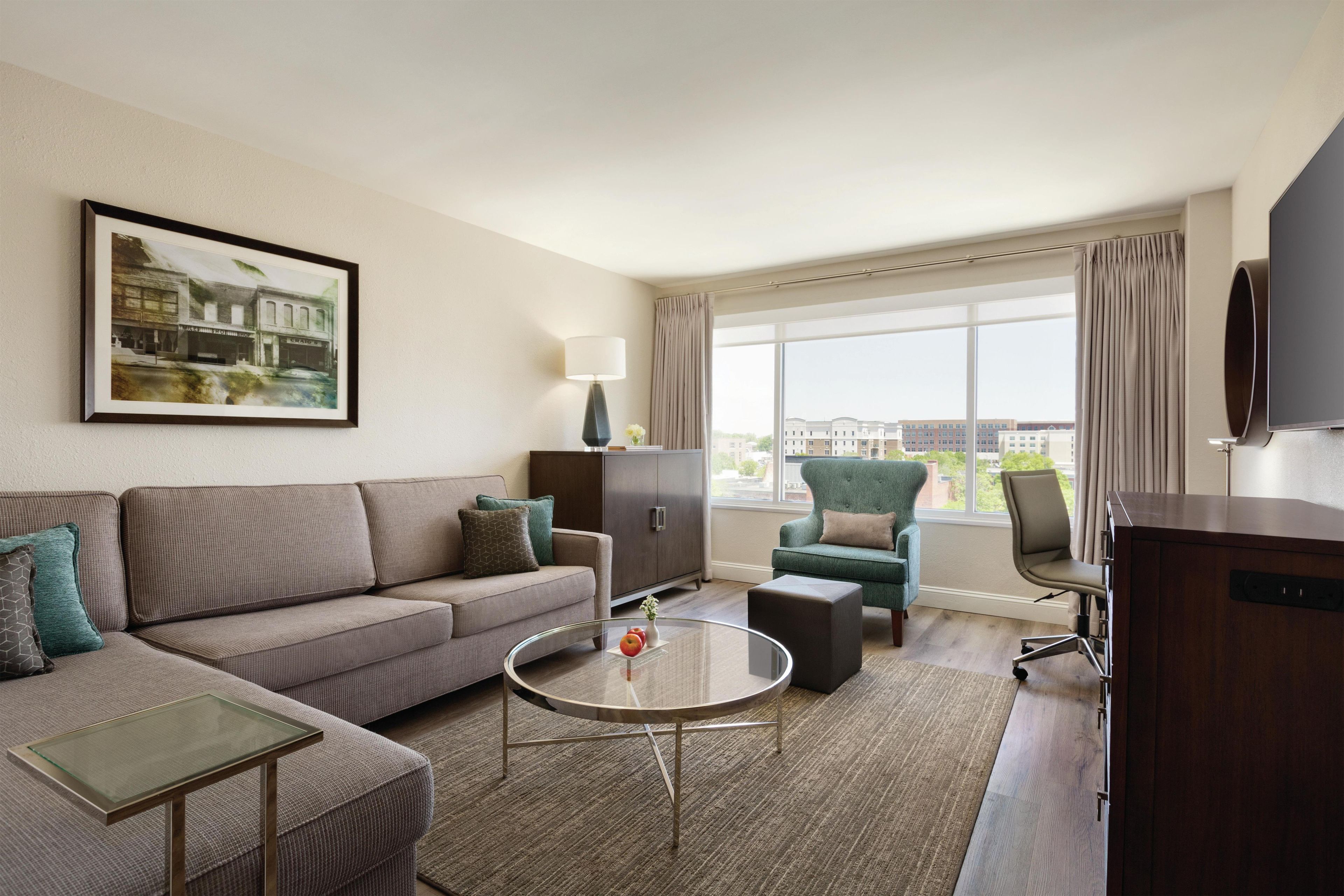 Une chambre d’hôtel moderne équipée d’un canapé, d’un fauteuil, d’une table basse et d’une grande fenêtre donnant sur la ville.