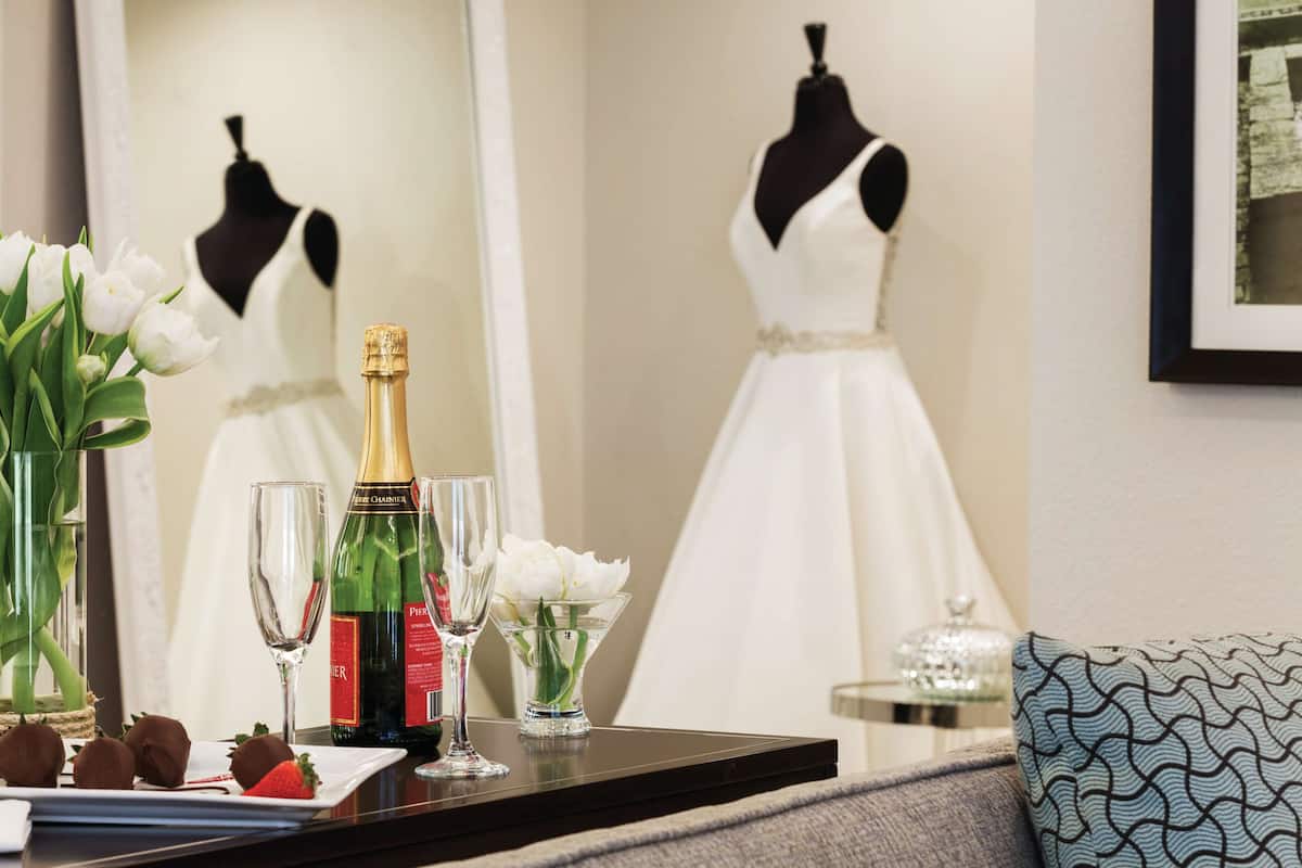 A table with champagne, glasses, flowers, and chocolates, with a dress on a mannequin in the background.