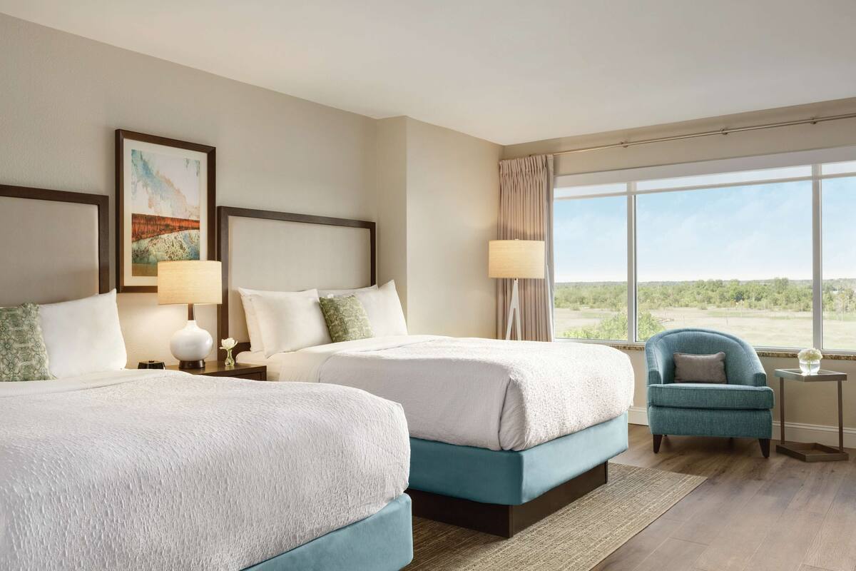 A hotel room with two beds, a chair, a lamp, and a large window with a view.