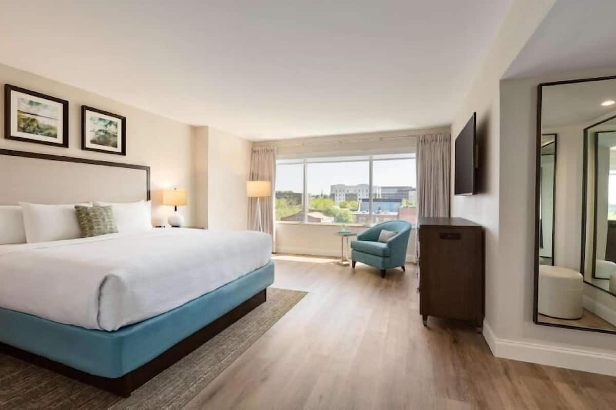 A hotel room with a large bed, a small table, a chair, a TV, and a window with a view of buildings.
