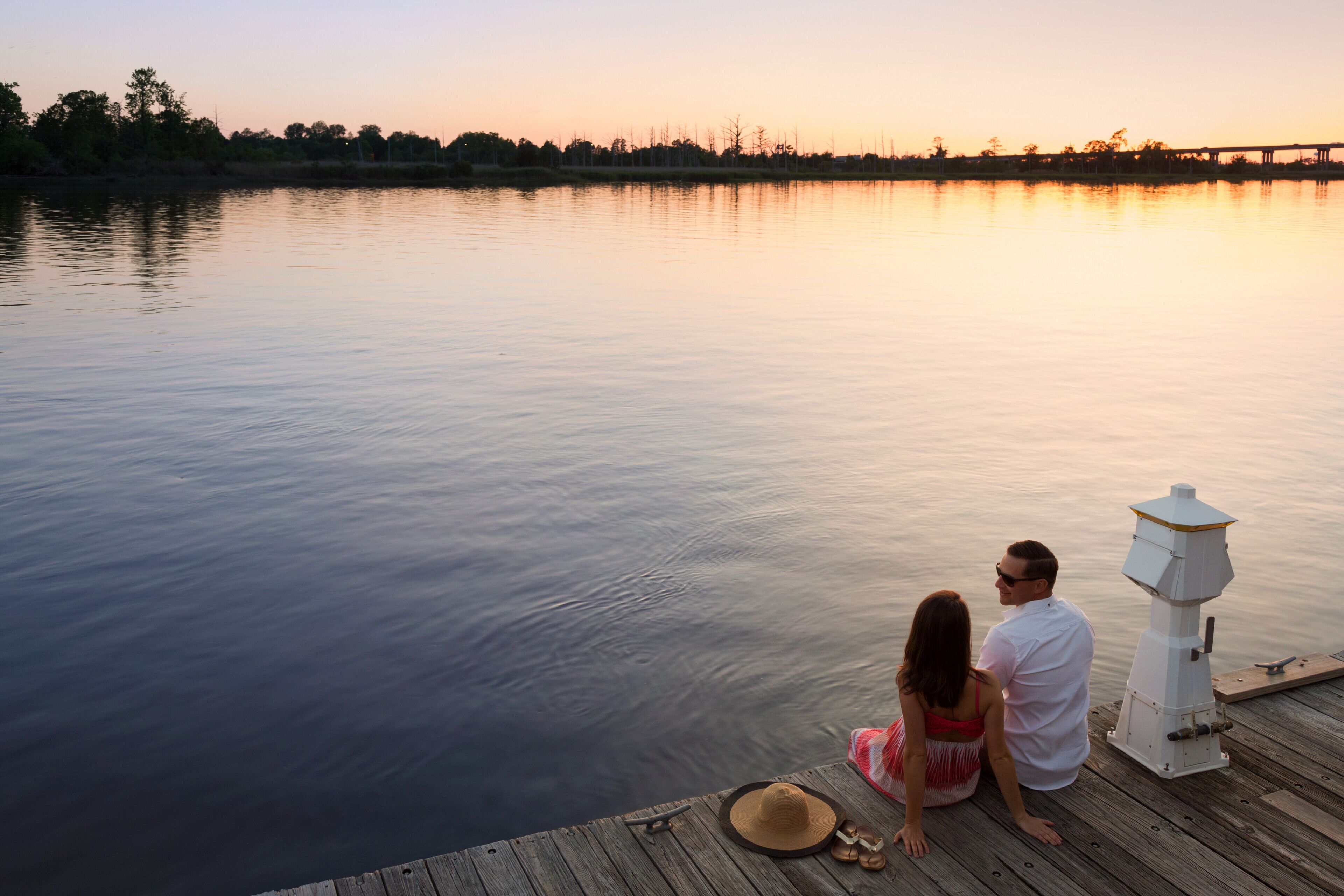 A couple sitting on a wooden dock overlooking a calm body of water with a sunset in the background.