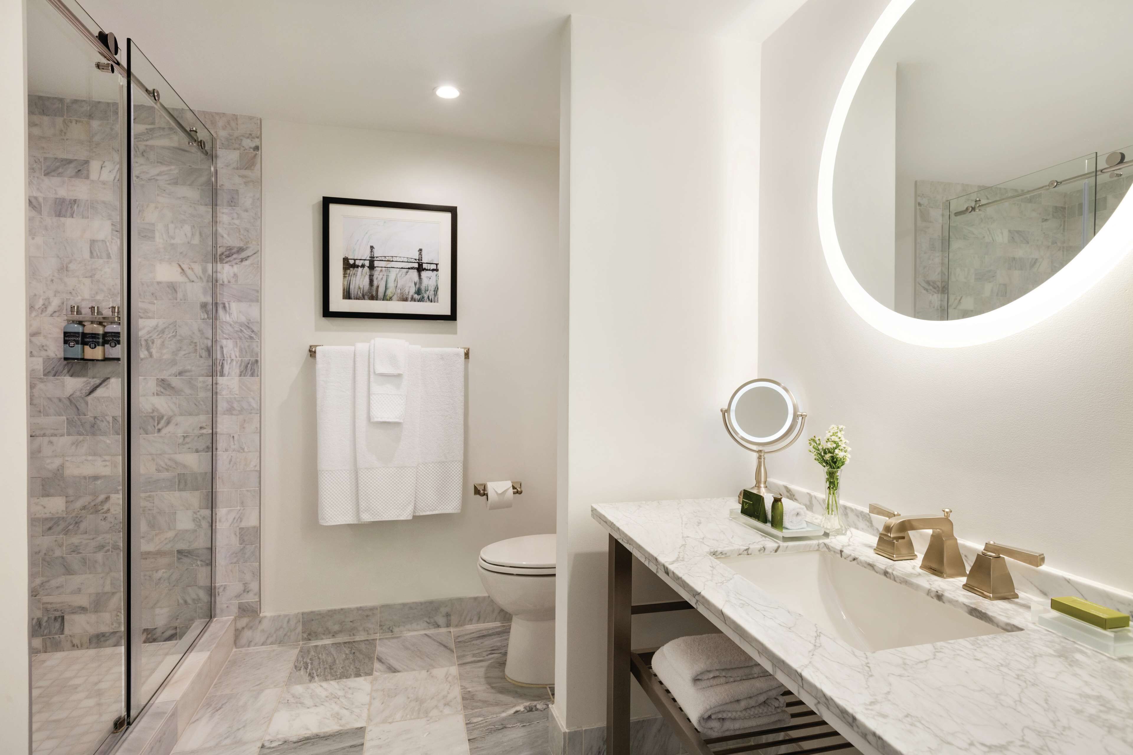 A modern bathroom with a glass shower enclosure, a marble-tiled floor, a toilet, a sink with a vanity, and a framed picture on the wall.
