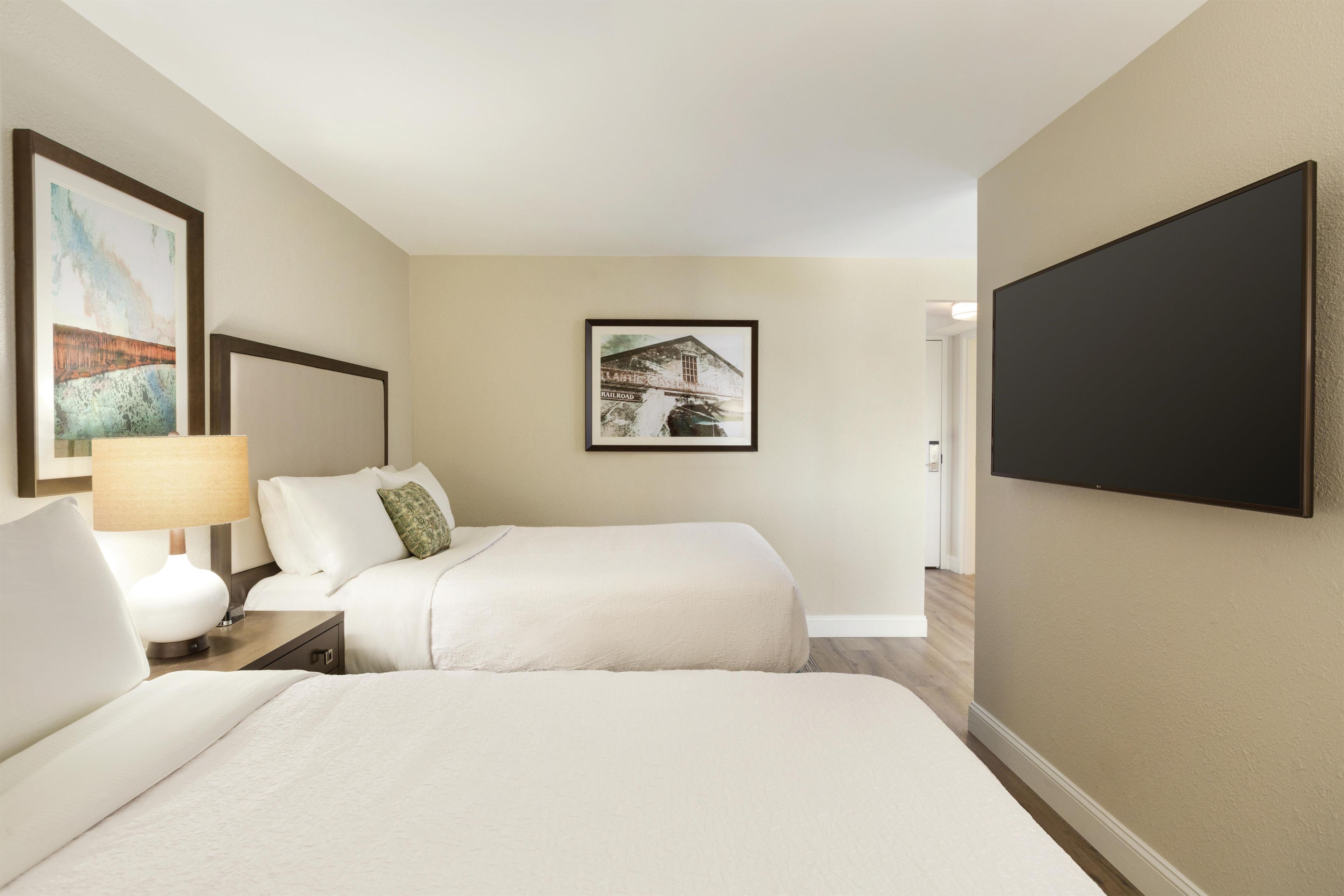 A hotel room with two beds, a nightstand, a lamp, and a wall-mounted TV.