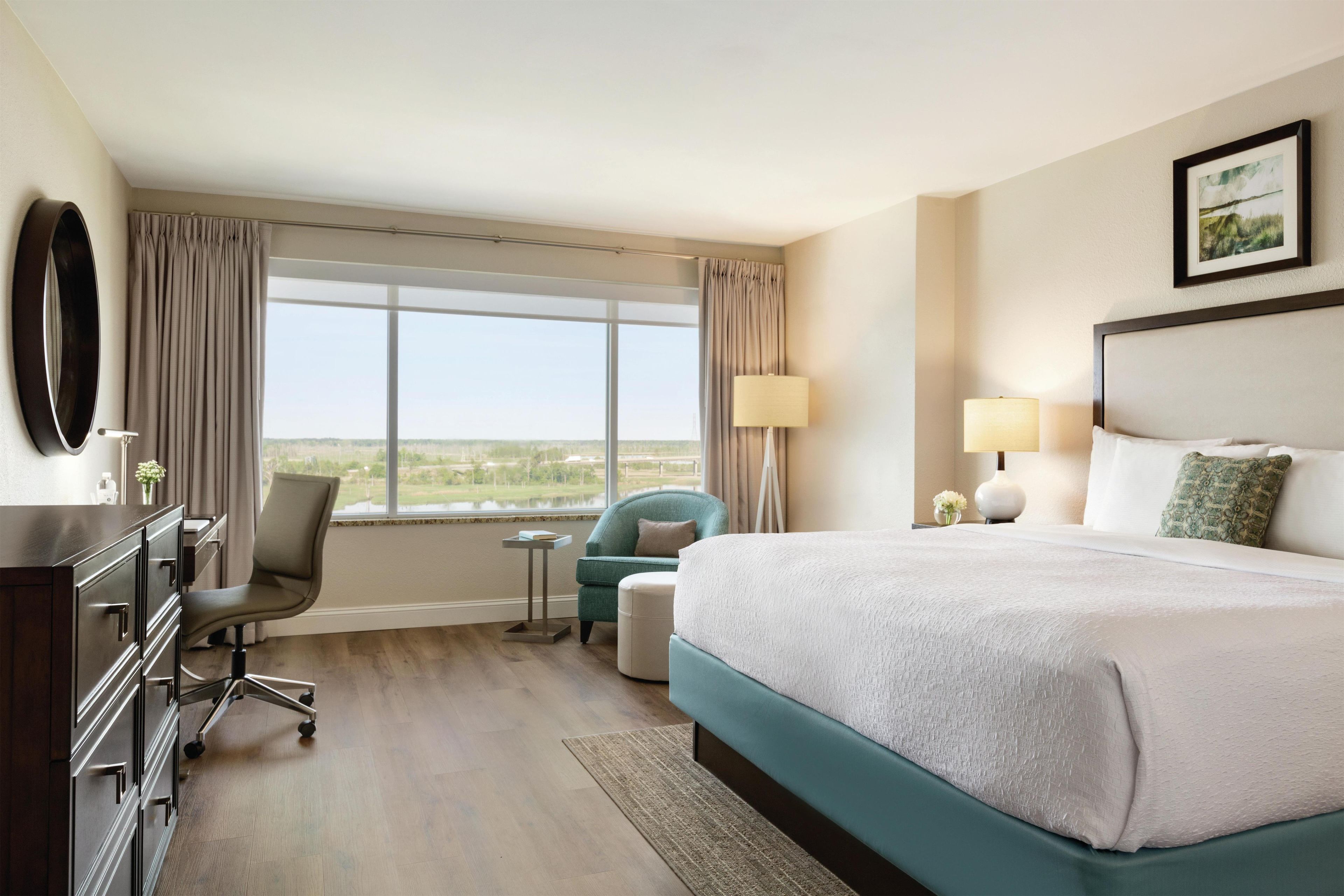 A hotel room with a large bed, a desk with a chair, a small table, and a view of a landscape through the window.