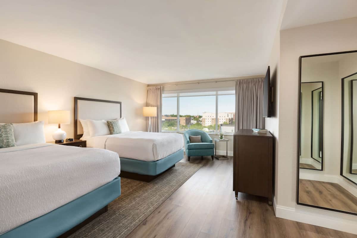 A hotel room with two beds, a desk, a chair, a TV, and a large window with curtains.