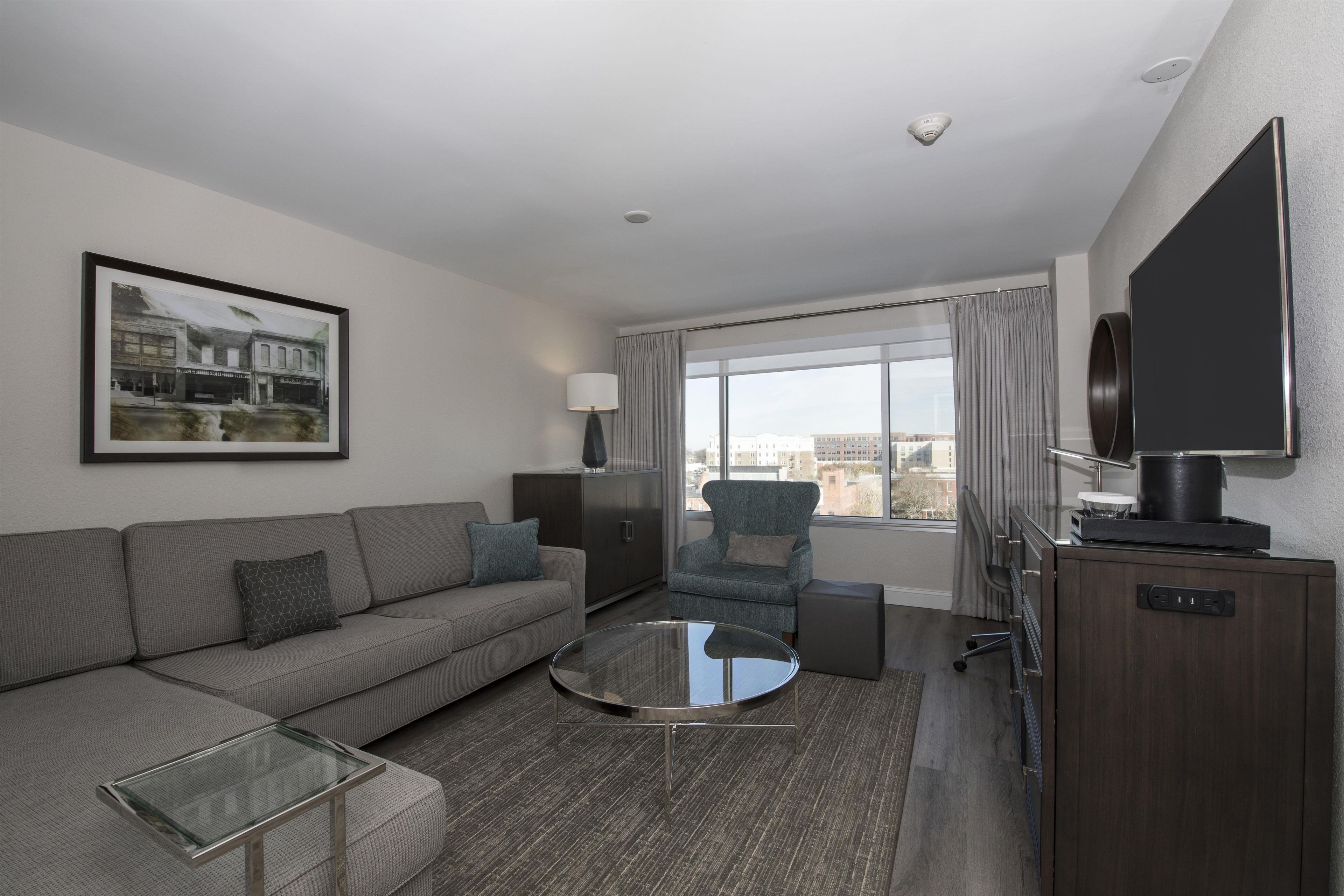 A modern hotel room with a sofa, armchair, glass coffee table, and a television.
