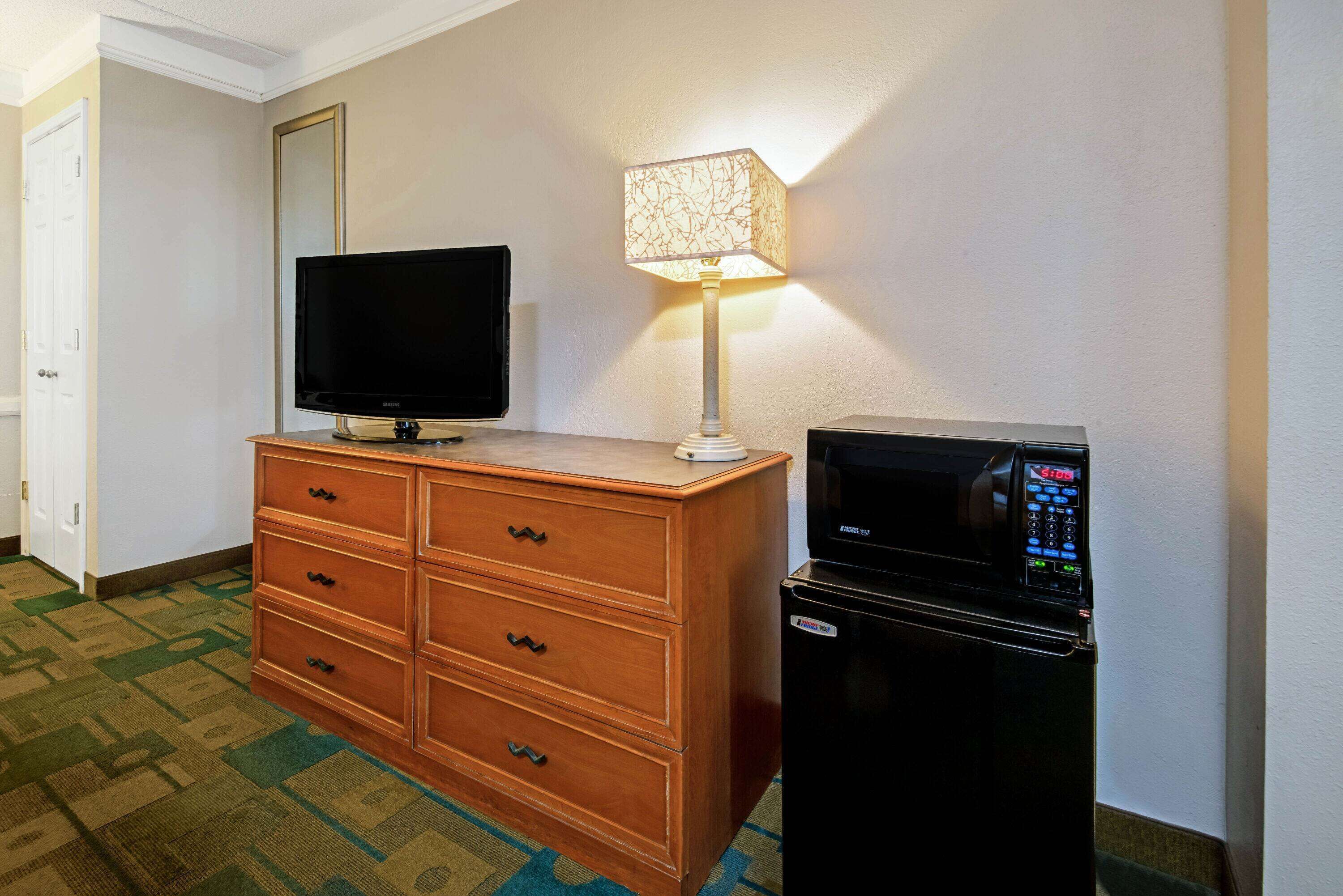 A hotel room with a wooden dresser, a flat-screen TV, a lamp, and a black refrigerator.
