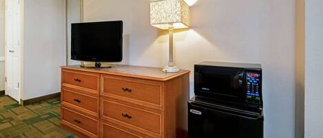 A hotel room with a wooden dresser, a flat-screen TV, a lamp, and a black refrigerator.