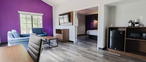 Habitación moderna con paredes moradas, piso de madera, una mesa de comedor, un sofá, una mesita, ventilador de techo, cama, televisor, microondas y cafetera.