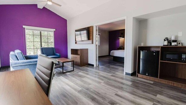 A modern room with purple walls, a wooden dining table, a blue sofa, a small wooden cabinet, and a kitchenette with a refrigerator and microwave. - La Quinta Inn by Wyndham Amarillo Mid-City (Amarillo)
