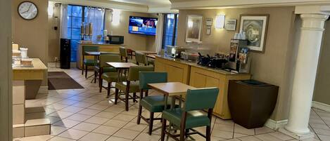 A hotel dining area with multiple tables and chairs, a kitchen area with a counter and appliances, and a television displaying a menu.