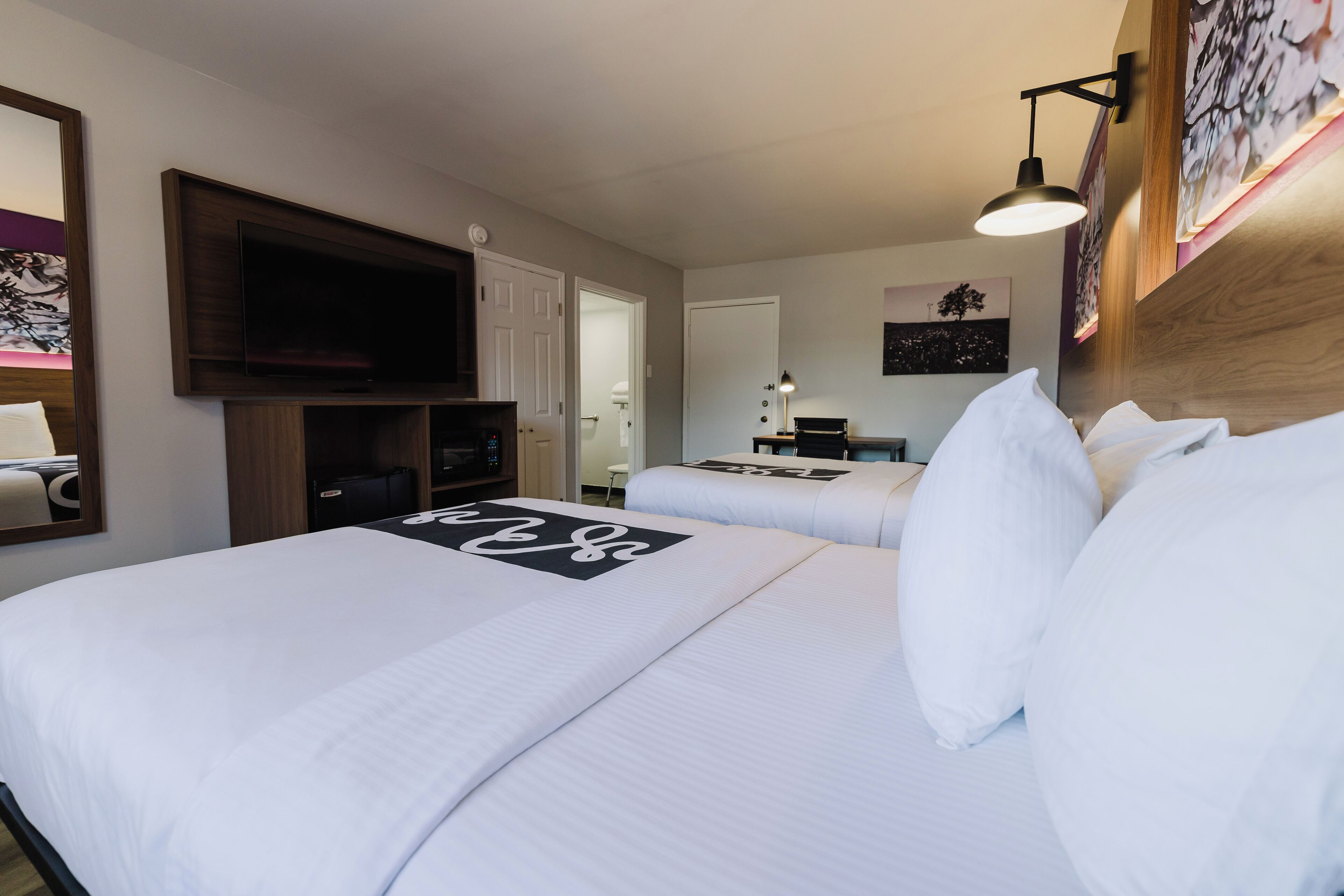 A hotel room with two beds, a TV mounted on the wall, a desk with a lamp, and artwork on the walls.