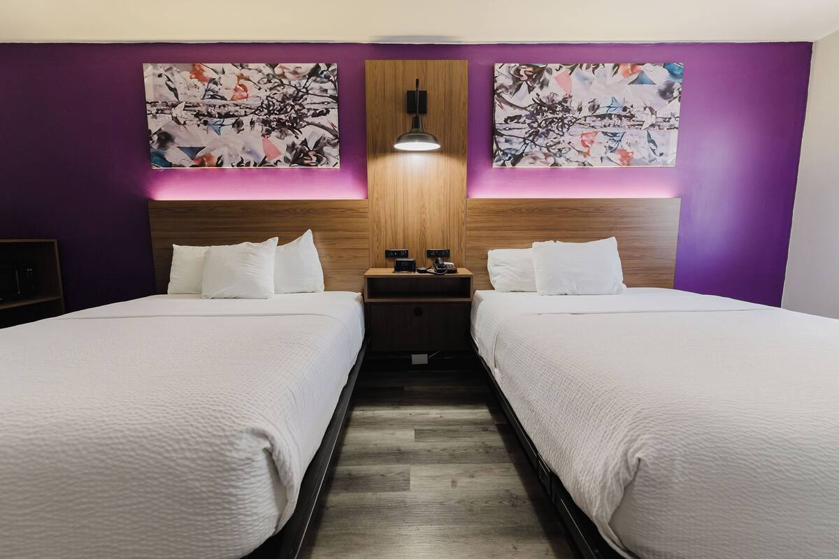 A hotel room with two beds, a wooden headboard, and two abstract paintings on the walls.