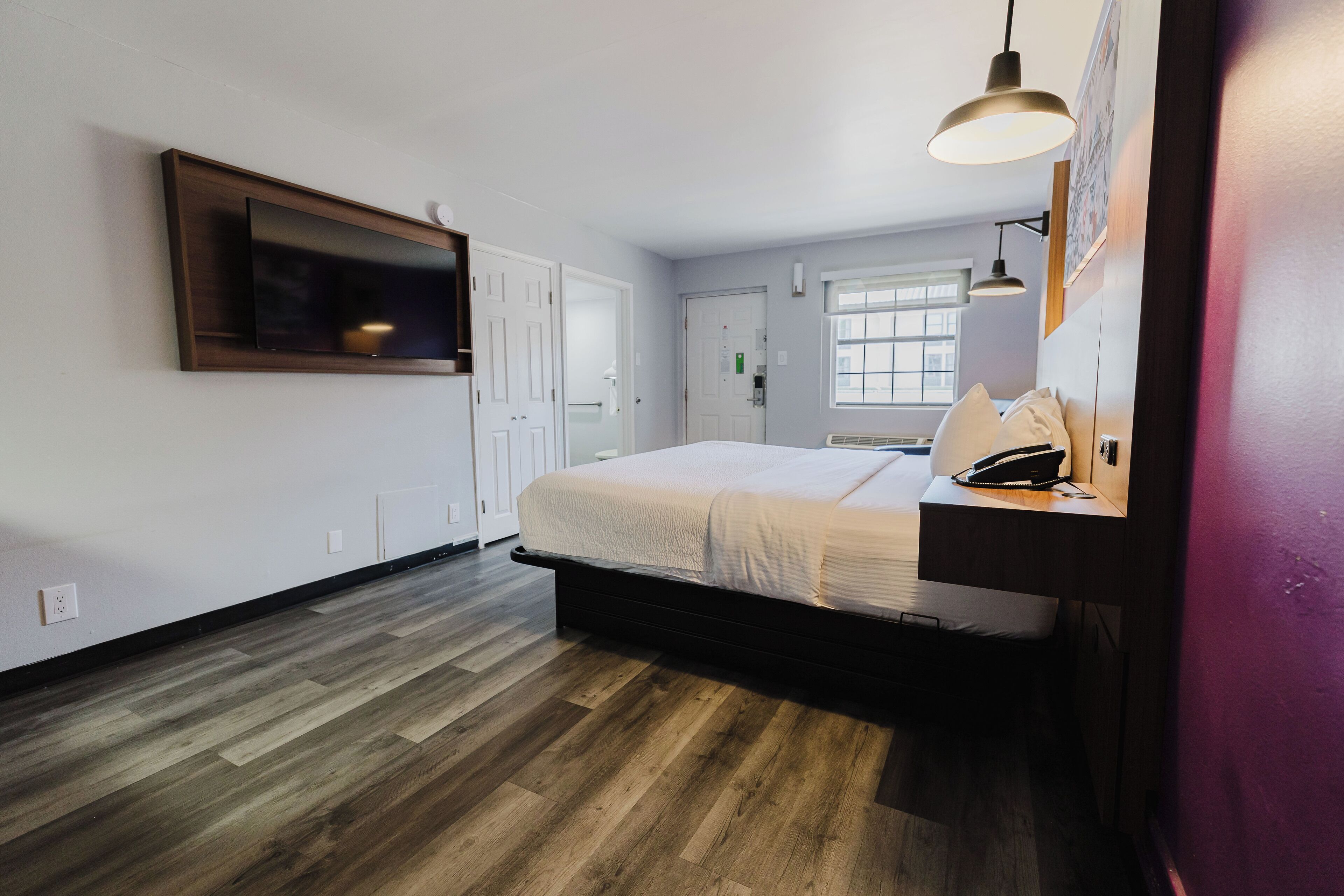 A bedroom with a bed, a mounted TV, a desk with a phone, and a window with blinds.