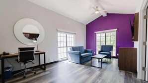 A modern living room with a purple accent wall, a round mirror, a blue sofa, a coffee table, and a desk with a chair. - La Quinta Inn by Wyndham Amarillo Mid-City (Amarillo)