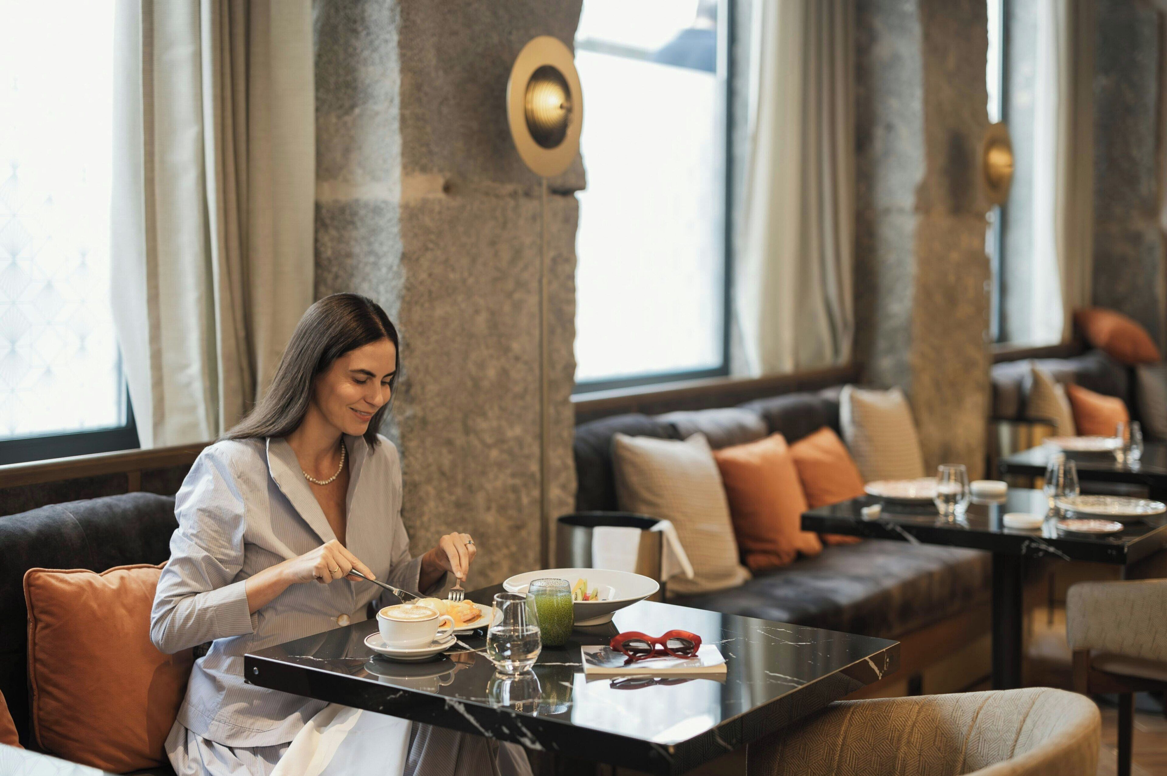 Una persona sentada a una mesa con comida, en una habitación con ventanas amplias y diseño interior moderno.