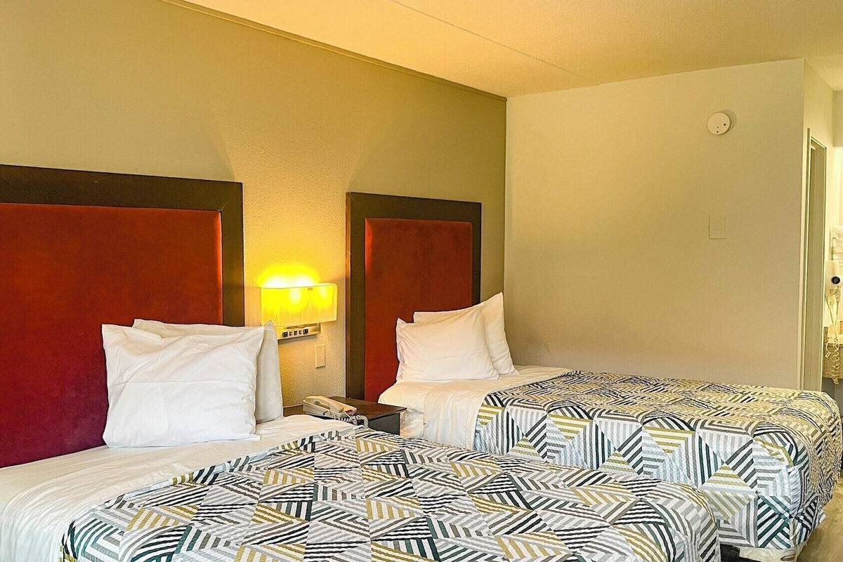 A hotel room with two beds, each with a patterned bedspread, a nightstand with a lamp, and a wall-mounted light fixture.