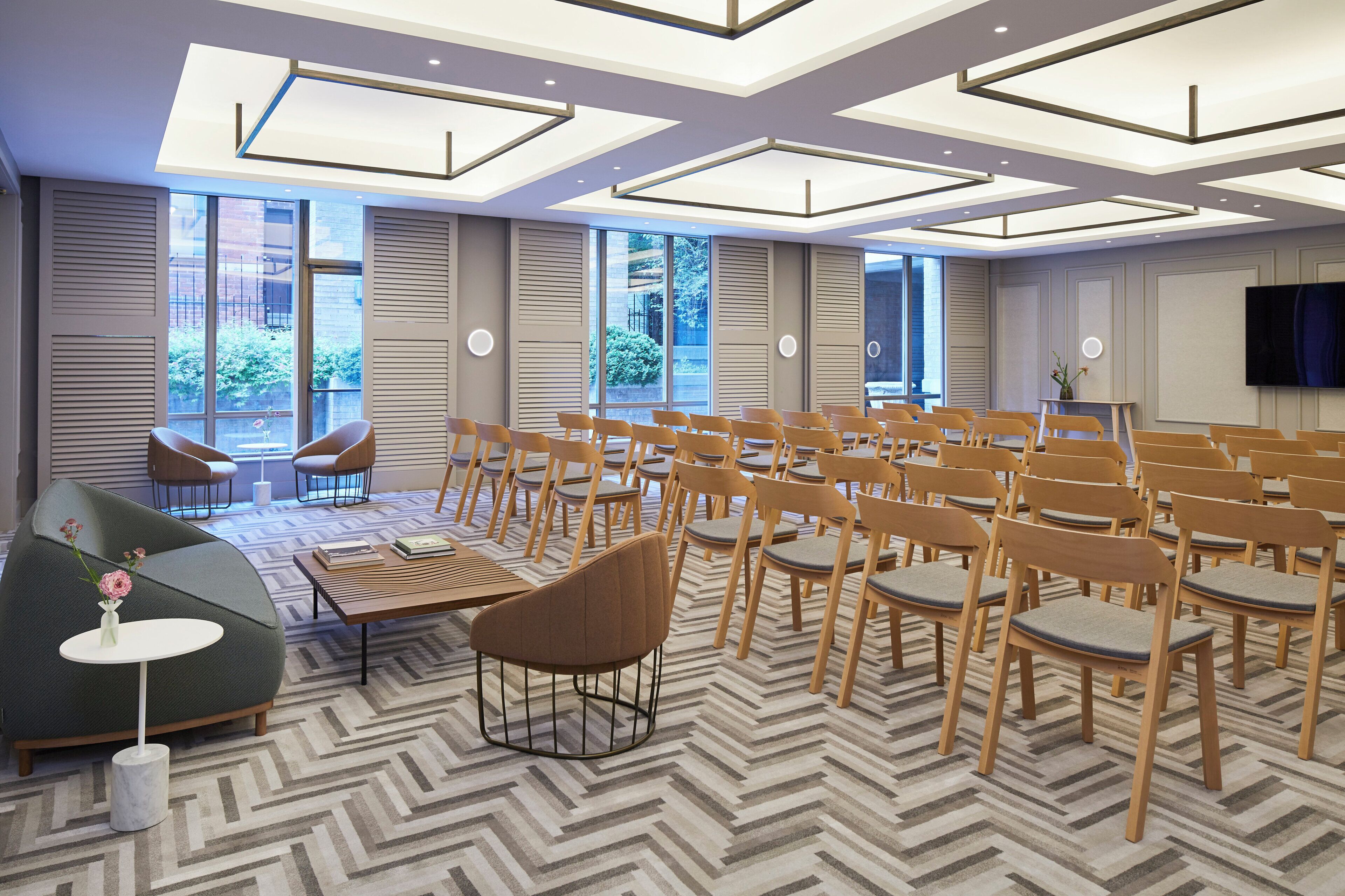 A conference room with a modern design, featuring a patterned carpet, wooden chairs, a sofa, and a television on the wall.