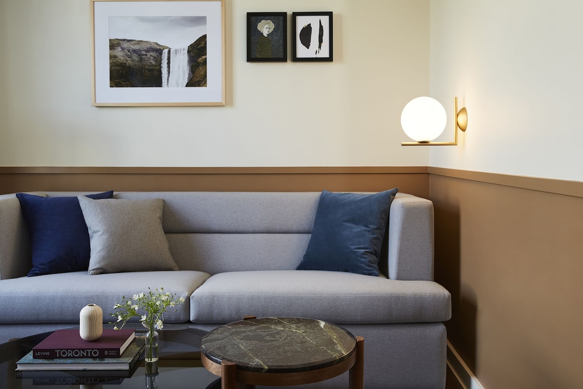 A modern living room with a grey sofa, blue cushions, a small round table with a vase of flowers, a book, and a framed picture of a waterfall.