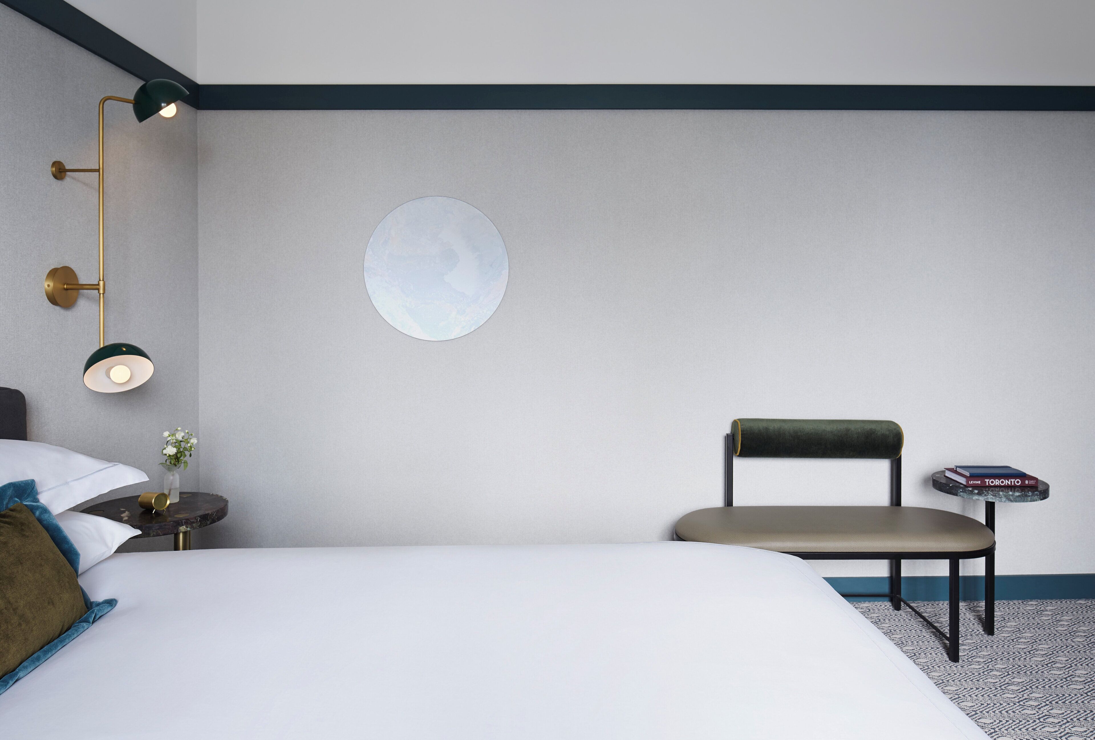 A hotel room with a bed, a round wall decoration, a chair, a small table with a book, and a nightstand with a lamp.
